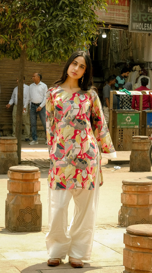 Woman in a patterned top and white pants standing on a street with people and shops in the background.