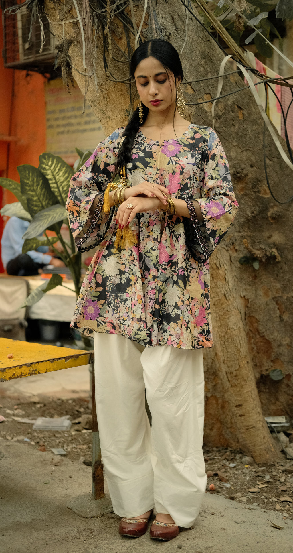 A woman stands outdoors wearing a loose, floral printed tunic top with wide sleeves and white wide-leg pants. The top has a multicolor floral pattern with pink, green, and blue tones on a dark base, paired with white pants and brown shoes.