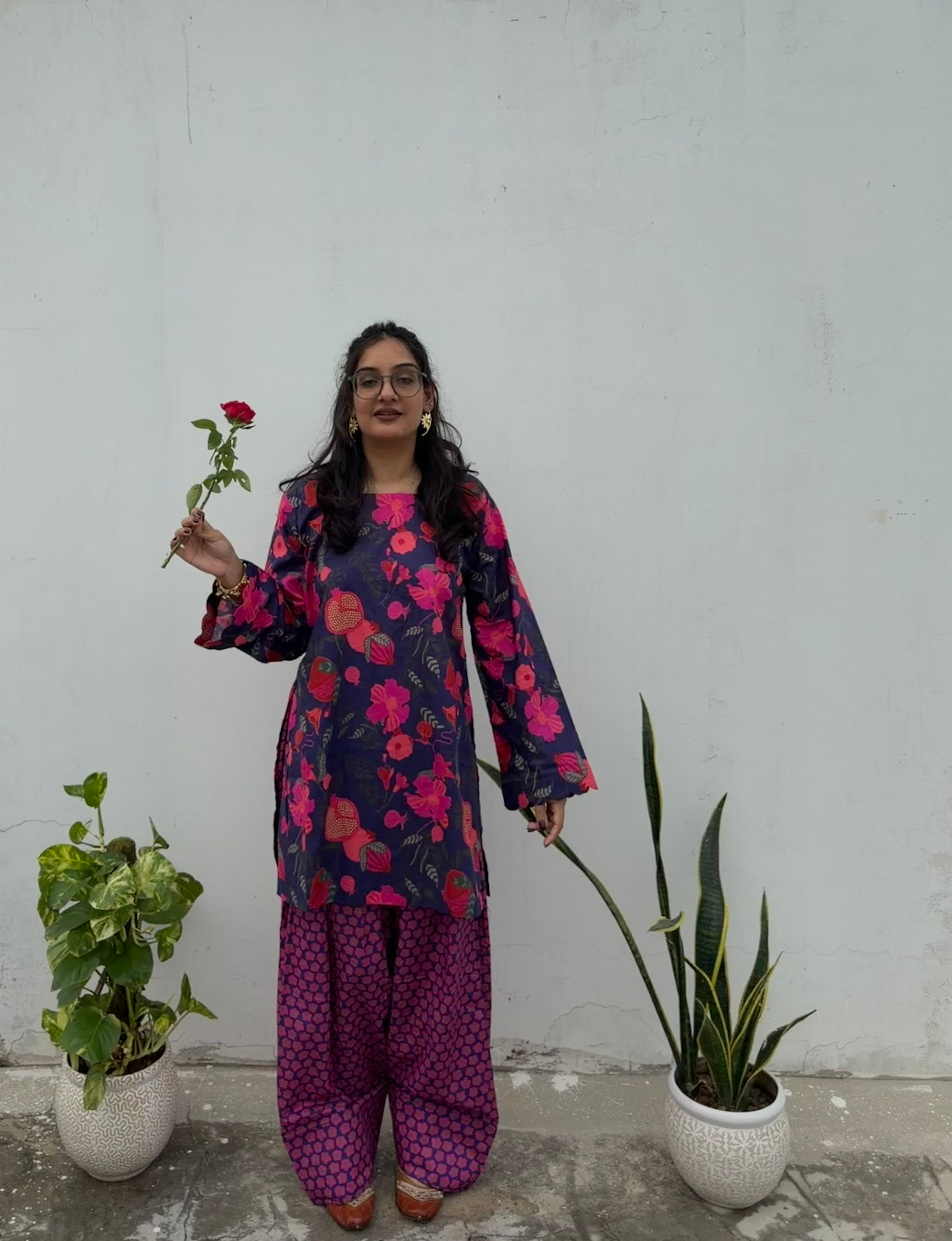 A woman wearing a dark navy floral kurta and matching patterned pants stands against a white wall, holding a single red rose. Two potted plants are on either side of her.
