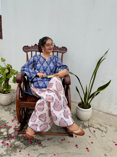 A woman wearing a blue and pink printed kurta set sits in a dark wooden rocking chair against a white wall, flanked by potted plants.