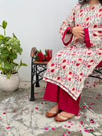 A woman seated on a black metal bench wearing a cream-colored printed kurta with red and pink floral motifs and deep pink wide-leg pants. A potted green plant and a small table with red roses are beside her against a plain light wall.