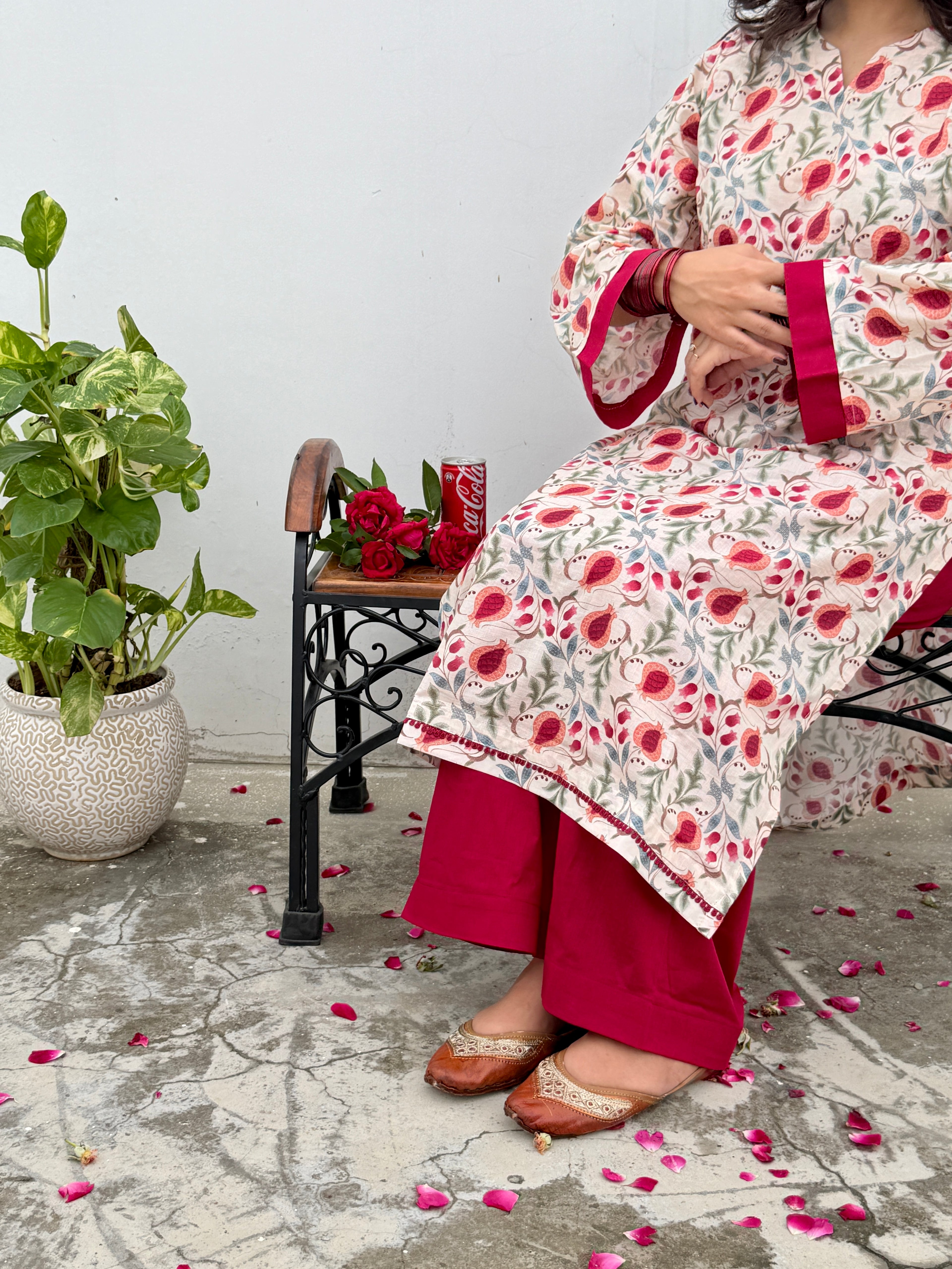 A woman seated on a black metal bench wearing a cream-colored printed kurta with red and pink floral motifs and deep pink wide-leg pants. A potted green plant and a small table with red roses are beside her against a plain light wall.