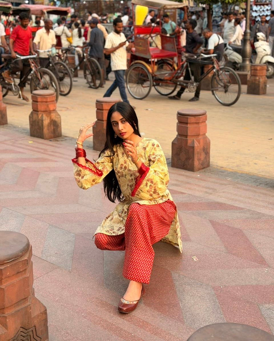 A woman posing in a traditional Indian outfit consisting of a beige floral kurta with red trim and red patterned pants, set against a busy outdoor street scene with rickshaws and pedestrians.